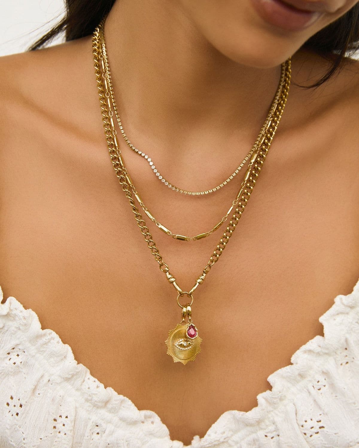 Close-up of a model's neckline in a white broderie top wearing three layered gold necklaces with a gemstone charm pendant