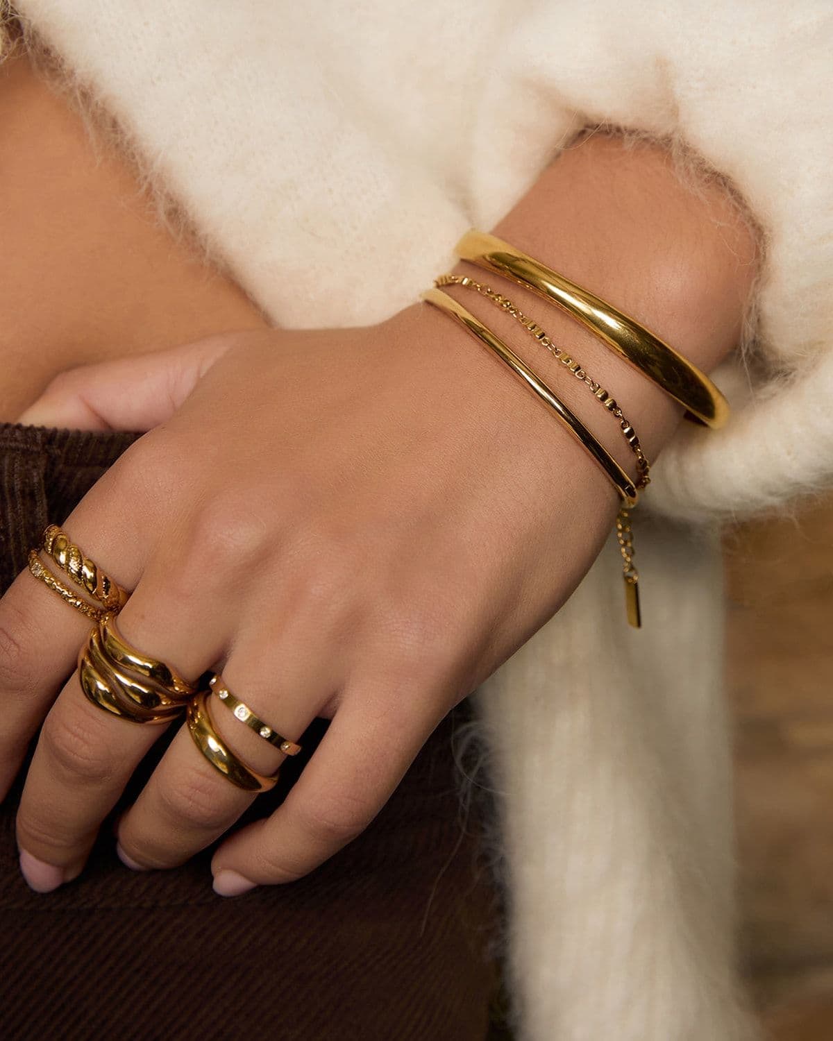 Close-up of a model's hand and wrist against a cream mohair sweater, stacked with gold bangles, chain bracelets and rings
