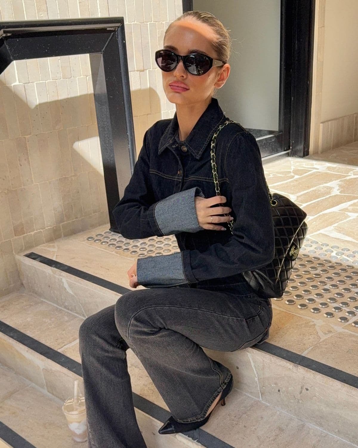 Model in a black denim jacket and flared jeans sits on travertine steps holding an iced coffee, quilted bag on her shoulder