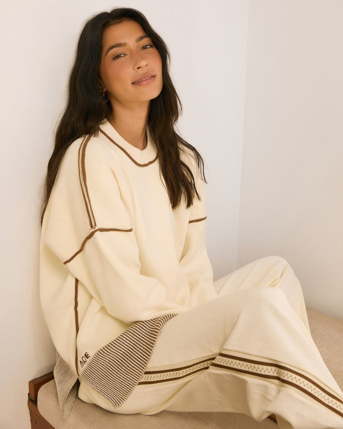 Model in an oversized cream ribbed-knit sweater and matching wide-leg pants seated against a plain wall, wearing a gold hoop earring and layered rings