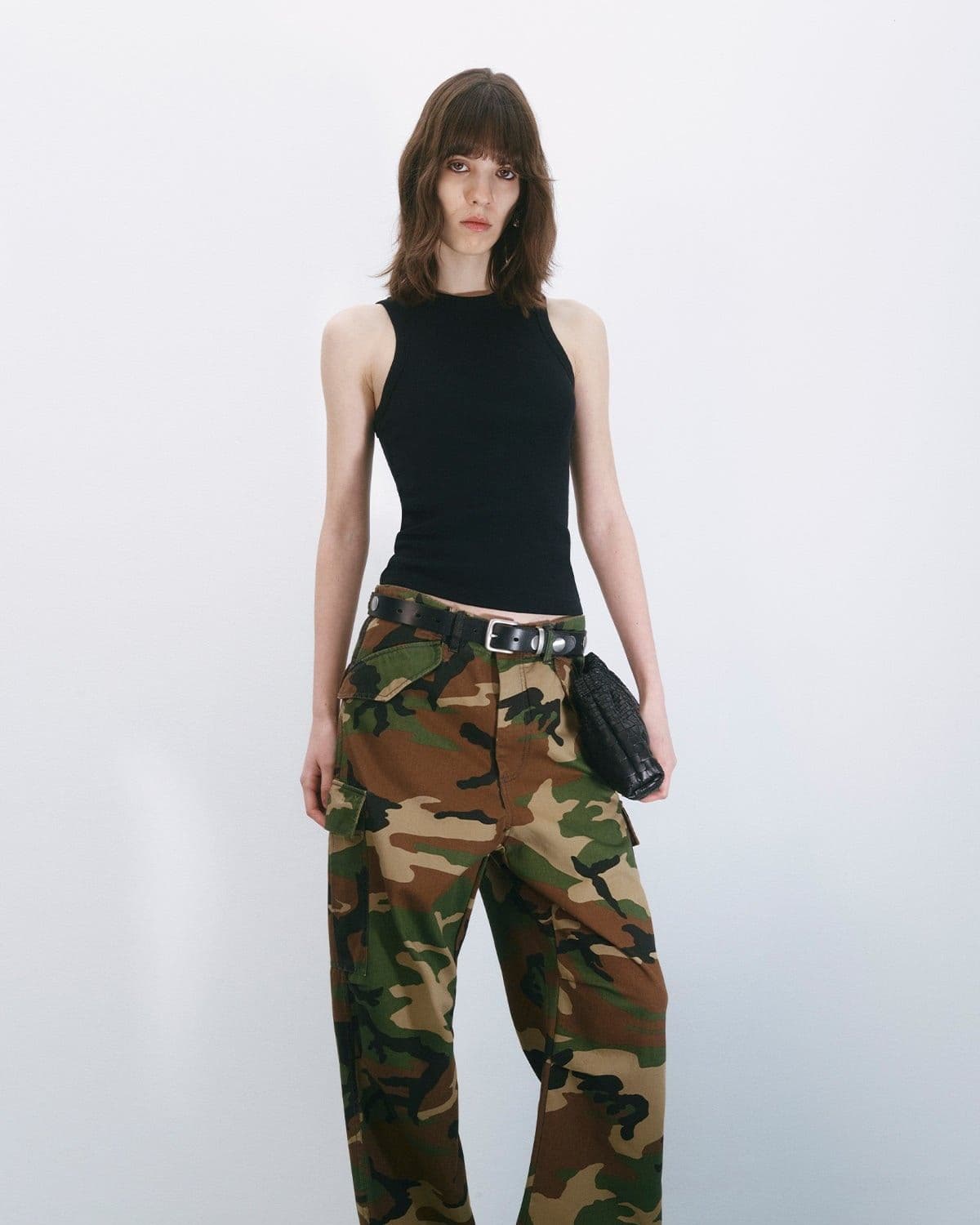 Model in camo cargo trousers, black tank top and belted mini bag, editorial studio lighting with cast shadow